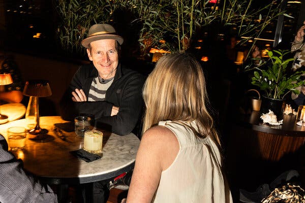 Denis O’Hare sits at a table with his arms crossed, smiling and wearing a brimmed hat. A woman sits at the same table with her back to the camera.