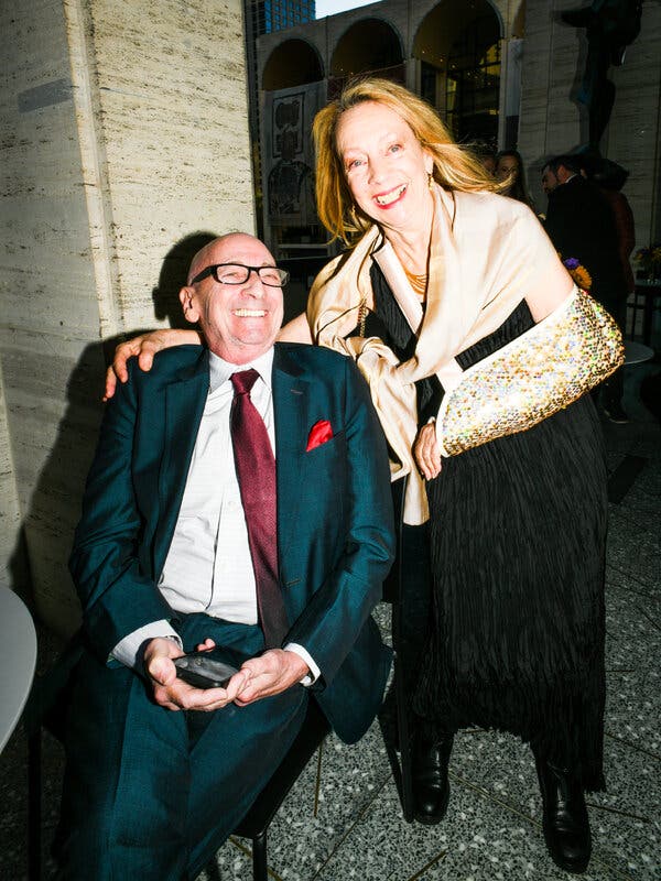 Alexander Bernstein, in a blue shirt and a red tie, sits smiling for the camera. Jamie Bernstein stands beside him in a black dress with her left arm in a glittery sling.