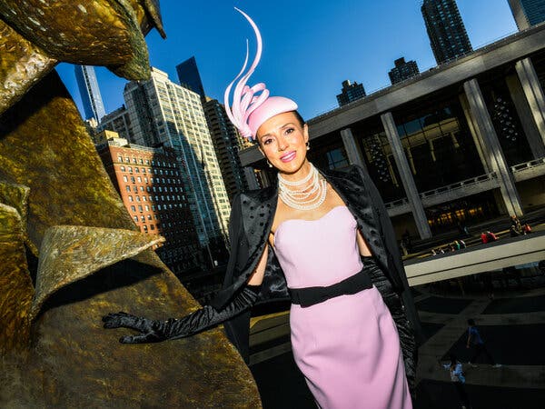 Genevieve Morritt, wearing a pink dress, black gloves, a pink hat with sculptural pink feathers on top, and a black jacket over her shoulders, stands outside David Geffen Hall.