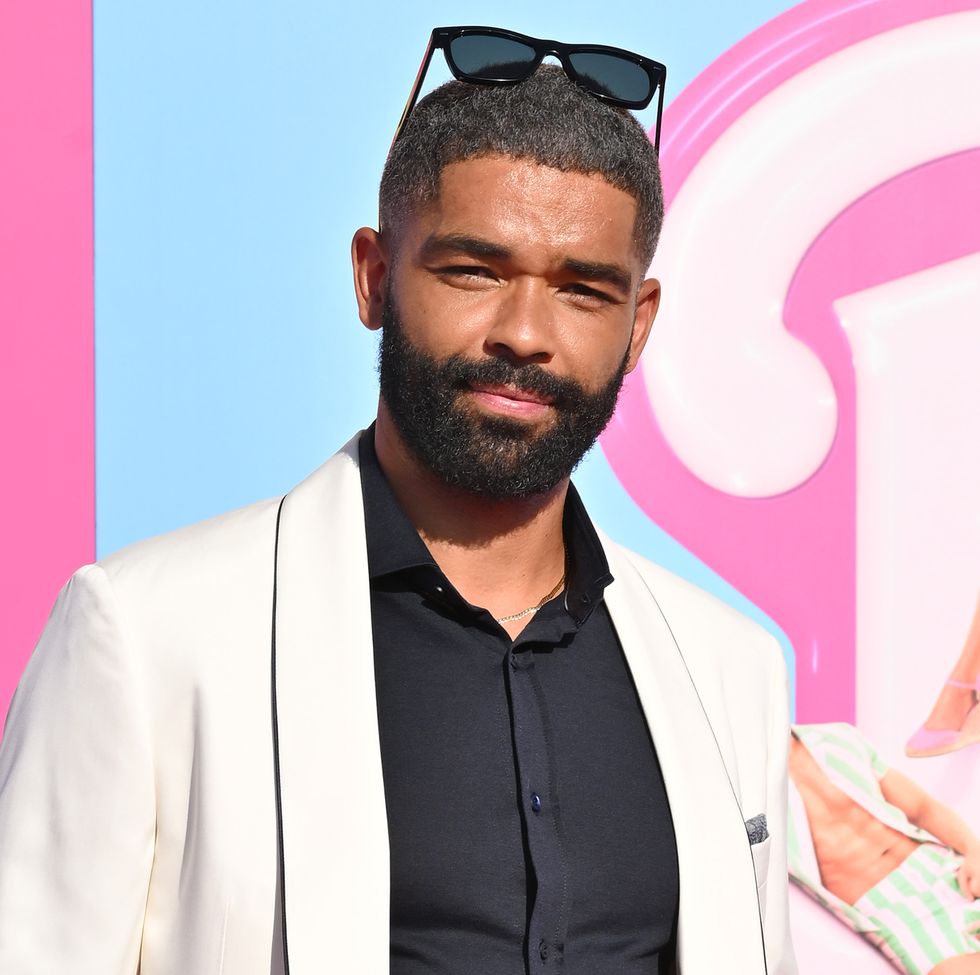 kingsley ben adir, a man with dark hair and a bear, wearing black pants a black shirt and a white suit jacket, on posing for the camera on a pink carpet at the premiere of the barbie movie in 2023