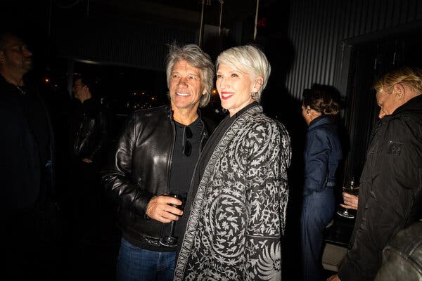 Jon Bon Jovi, wearing a leather jacket over a black T-shirt, and Maye Musk, wearing a patterned black-and-white jacket, stand together at a party, smiling.