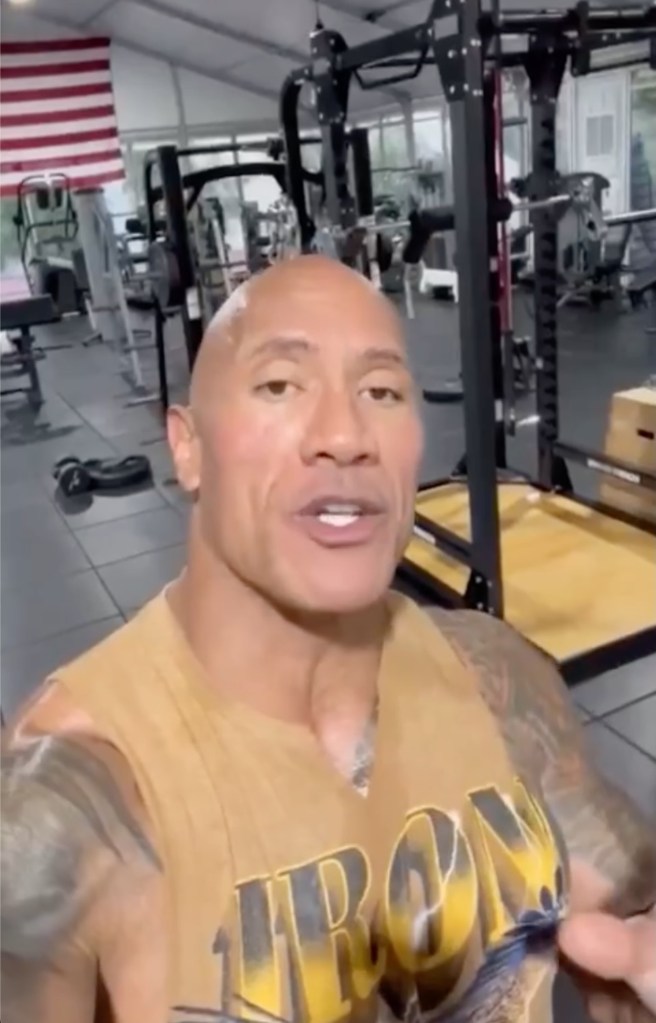 Dwayne "The Rock" Johnson in a mustard colored sleeveless shirt with the word iron on it, in the gym, holding up the camera and looking into it