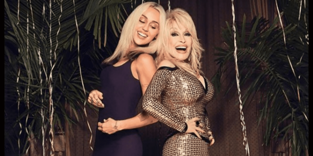 Two women posing together, one in a purple dress and the other in a Dolly Parton-inspired checkered outfit, standing among large potted plants with joyful expressions.