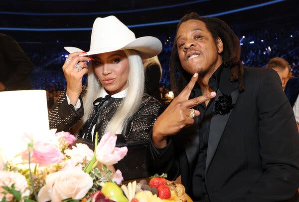 Beyoncé and Jay-Z pose at a table set with flowers at the Grammy awards ceremony. She wears a white cowboy hat; he’s in a black tux with a black shirt, sans tie. 