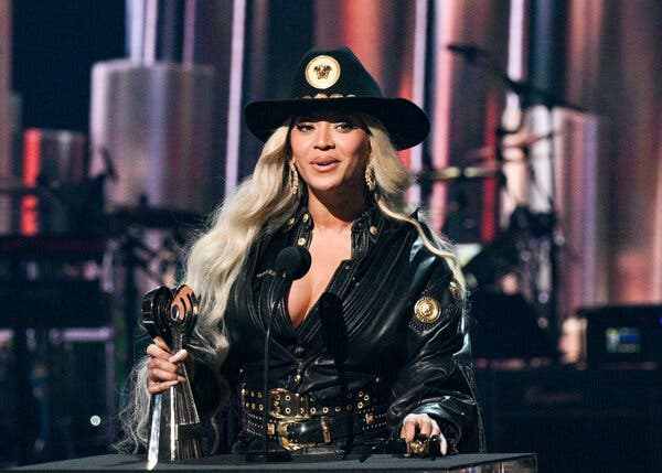 Beyoncé stands at a podium accepting an award. Her oversize leather jacket is embellished with gold details. She wears a black cowboy hat atop her very long platinum hair. 