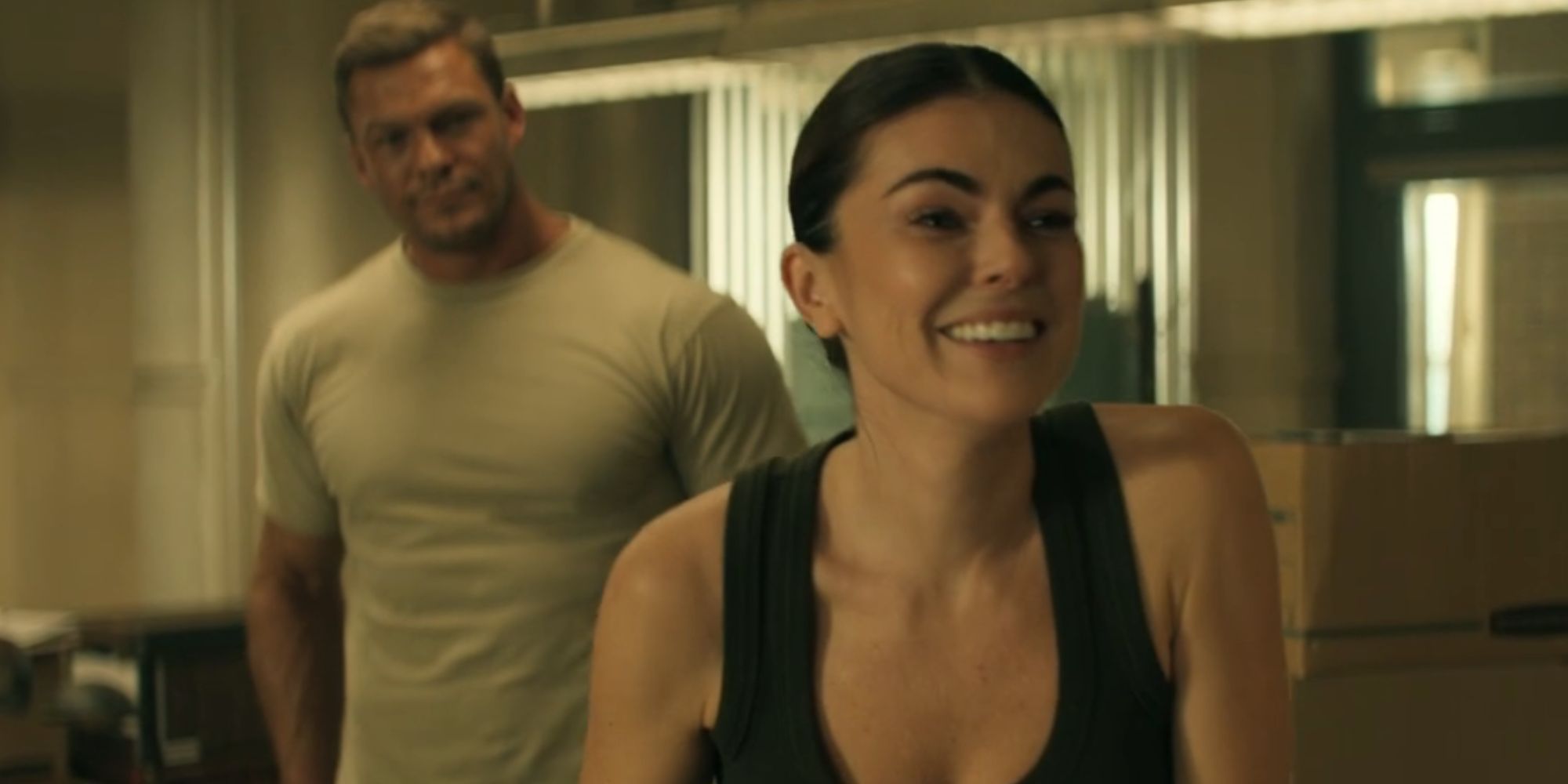 Serinda Swan smiling as Karla Dixon and Alan Ritchson's Reacher standing behind her in Reacher