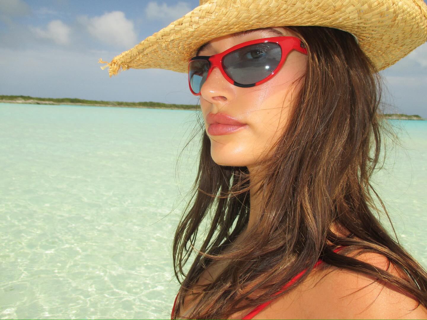 Hailey Bieber wears sunglasses and a straw hat on a beach.